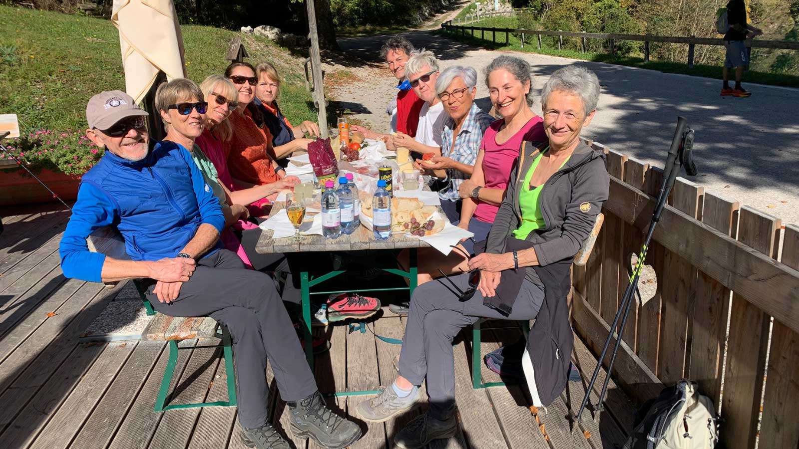 Gruppe von 10 Personen sitzt in Wanderkleidung zur Brotzeit an einem langen Holztisch auf zwei Bänken, vor sich Wasserflaschen, Weintrauben, Käse, Brot. Im Hintergrund die Bergstraße