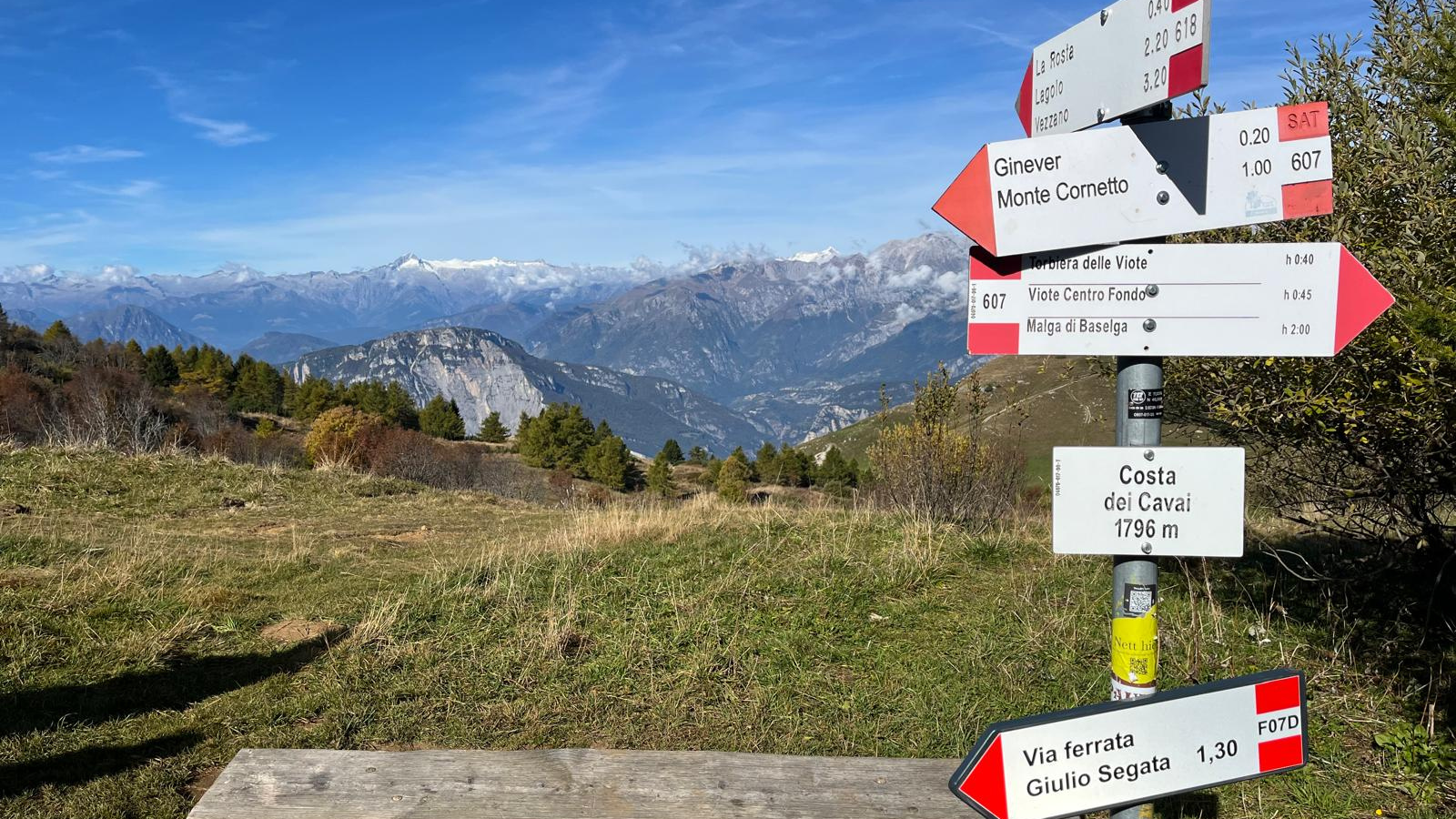 Blick von einer Grasebene in die teilweise schon schneebedeckten felsigen Gipfel der Brenta Dolomiten. Im Vordergrund mittig eine Bank und daneben vier Wegweiser die in unterschiedliche Richtungen weisen sowie die Angabe der aktuellen Höhe mit 1796 m