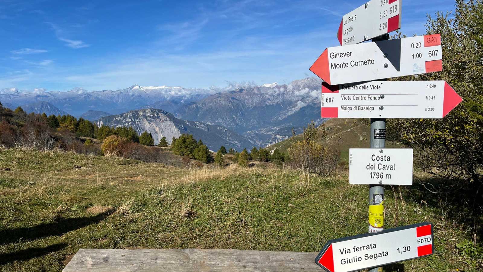 Blick von einer Grasebene in die teilweise schon schneebedeckten felsigen Gipfel der Brenta Dolomiten. Im Vordergrund mittig eine Bank und daneben vier Wegweiser die in unterschiedliche Richtungen weisen sowie die Angabe der aktuellen Höhe mit 1796 m