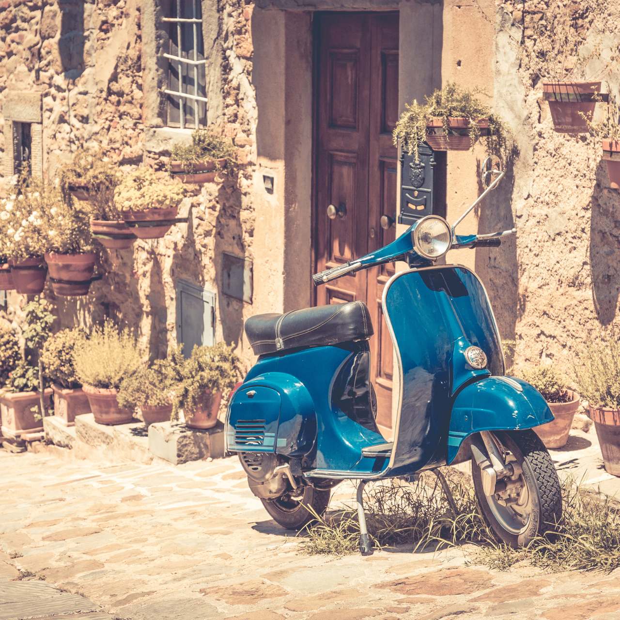 blaue Vespa vor einer steinernen Häuserreihe in der Toskana mit verdörrten Kräutertöpfen an der Hauswand