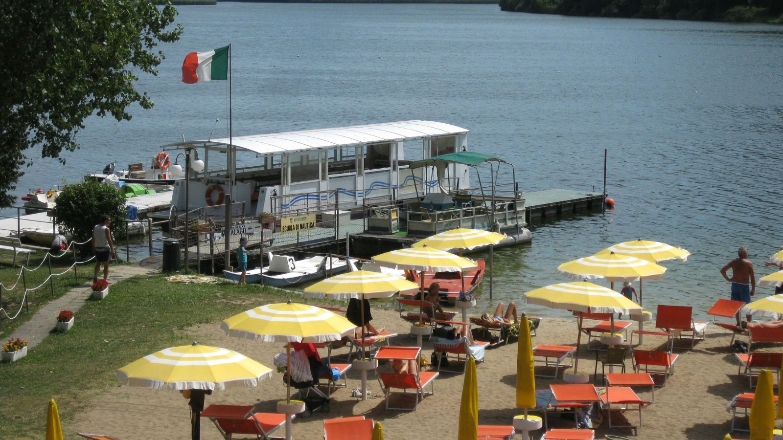 See-Anlegestelle mit Strandbad, aufgespannten Sonnenschirmen, roten Liegestühlen und Leute beim Sonnenbad. Es weht die italienische Flagge